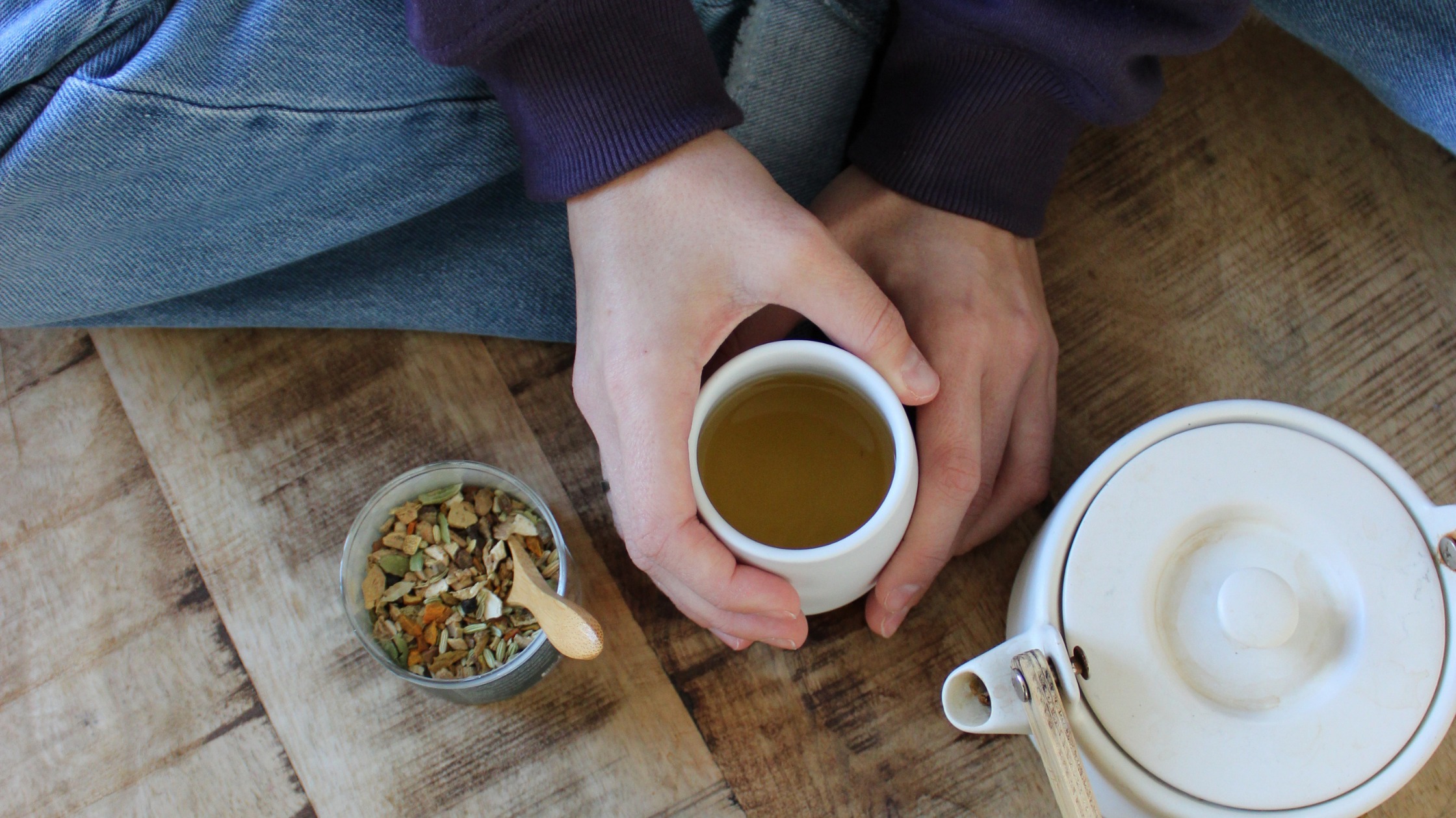 Hands holding a small white cup of herbal tea on a wooden table, with a white teapot and a jar of loose herbs nearby.